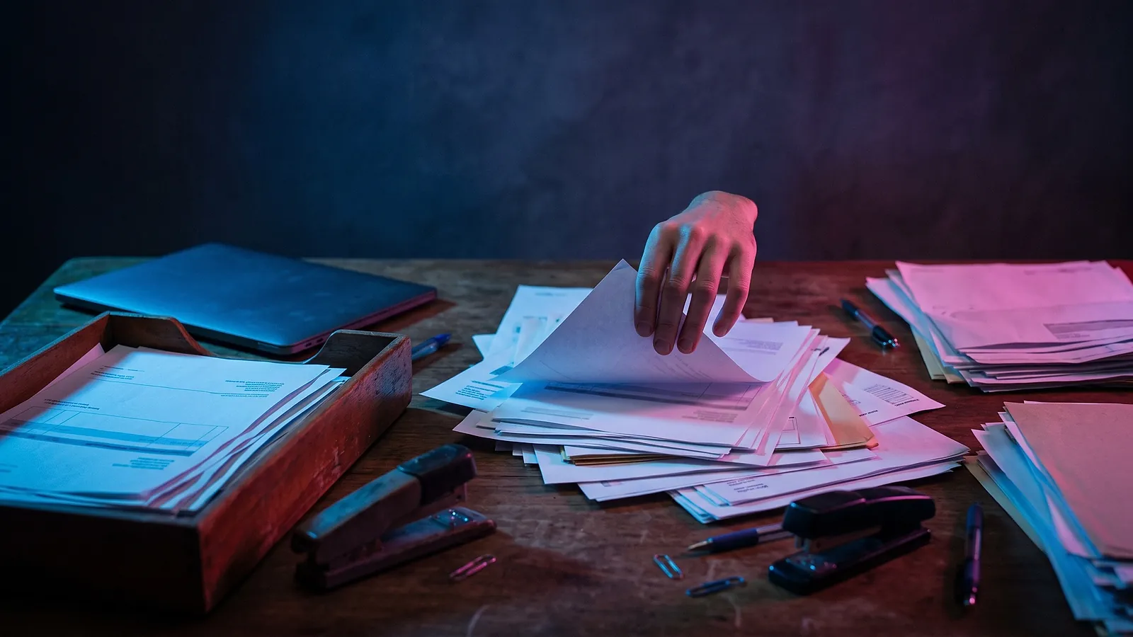 Une main trie une pile de documents papier débordant d'une bannette sur un bureau en bois, baigné de lumière bleue et rose.